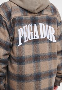 Brązowa i niebieska koszula flanelowa w kratę z dużym białym napisem "PEGADOR" na plecach. Posiada luźny krój i miękką teksturę.