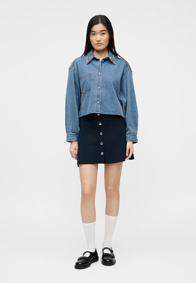 Νεαρή γυναίκα που φοράει κοντό τζιν πουκάμισο με διακοσμητικό γιακά, μίνι φούστα σε ναυτικό μπλε με κουμπιά, λευκές κάλτσες και μαύρα μοκασίνια, στέκεται όρθια.
