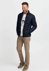 Svart quiltet jakke med høy krage og glidelås, kombinert med en grå t-skjorte og beige chinos, fullført med brune semskede sko.