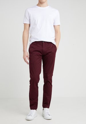 Homme portant un t-shirt blanc uni à manches courtes, un pantalon bordeaux et des baskets blanches, debout avec une main dans la poche, devant un fond neutre.