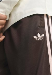 Brązowe spodnie sportowe z różowymi paskami po bokach; wyszywane białe logo Adidas na lewym udzie; gładka tekstura i luźny fason.
