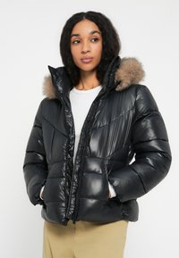 Frau in einer schwarzen, glänzenden Steppjacke mit Kapuze aus Fell, weißem Shirt und beigen Hosen, die mit den Händen in den Taschen der Jacke leicht zur Seite schaut.