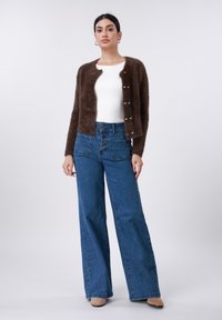 Pruun karvane kardigan, millel on nööpidega sulgemine, kombineeritud kõrge vöökohaga siniste denimist laiade pükstega. Allosas valge liibuv särk, neutraalsed kingad.