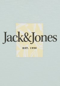 Svijetloplavi materijal s velikim žutim kvadratom i bijelim apstraktnim uzorkom, prikazuje tekst "Jack & Jones EST. 1990" u podebljanim crnim slovima.