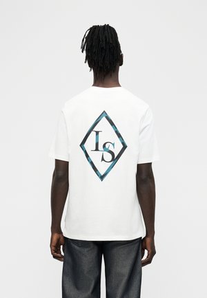 Muž s krátkymi dredmi, oblečený v bielom tričku s modro-čiernym diamantovým logom na chrbte obsahujúcim písmená "LS".