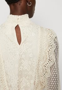 Krämfärgad spetsblus med blommönster, volangkrage, nyckelhål i ryggen och texturerade ärmar med intrikata vävda detaljer.