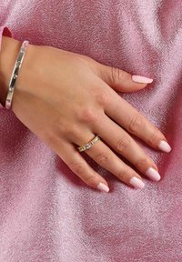 Silverarmband med klara stenar och guld ring med alternerande klara och färgade stenar på en hand mot en texturerad rosa bakgrund.