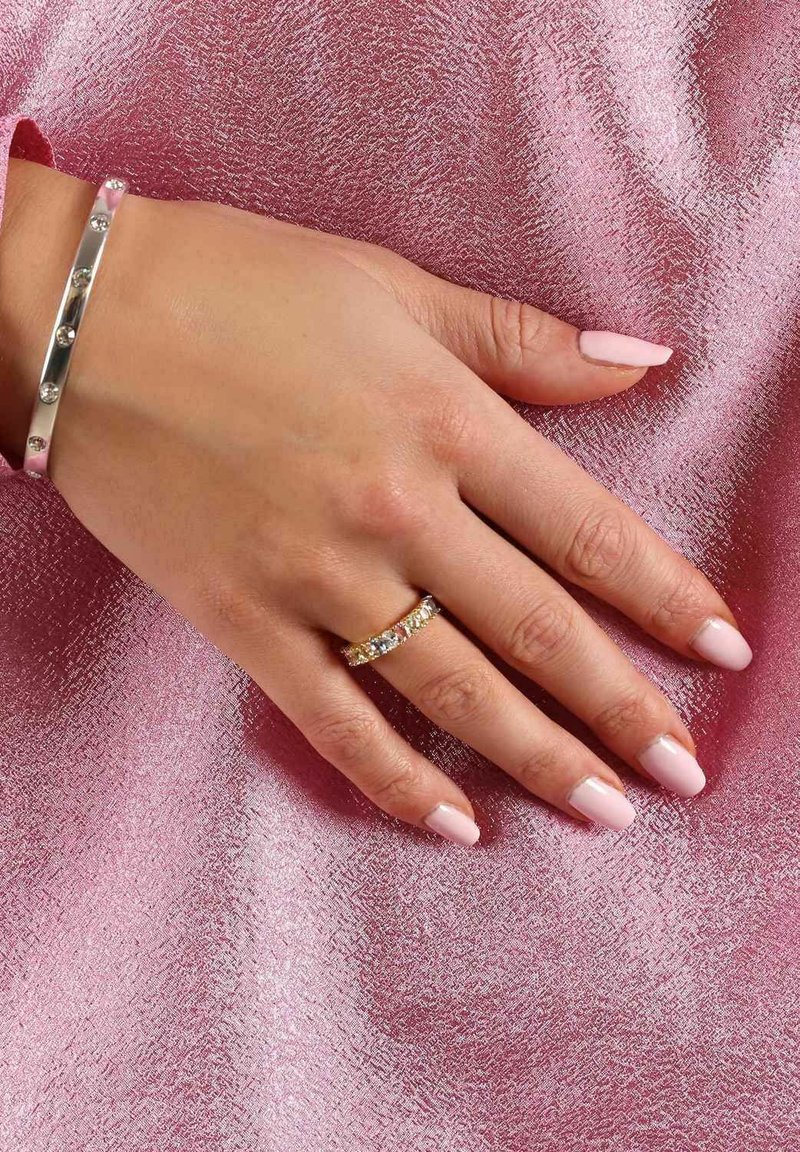 Silverarmband med klara stenar och guld ring med alternerande klara och färgade stenar på en hand mot en texturerad rosa bakgrund.