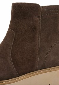 Bottine en daim marron avec semelle beige et couture visible, présentant une surface légèrement texturée et un détail de marque près du bord supérieur.