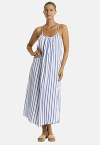 Un modello posa in un abito maxi senza maniche, a righe blu e bianche, indossando sandali beige, su uno sfondo neutro e con un'espressione sicura.
