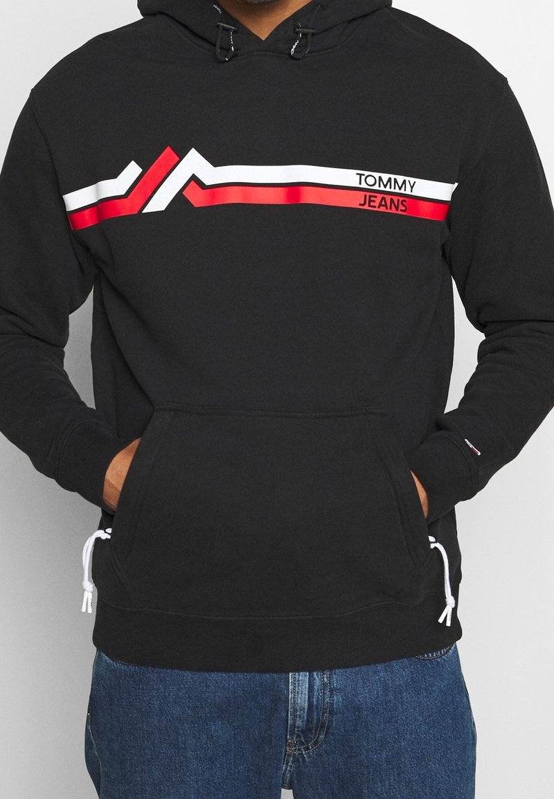 Persona che indossa un hoodie nero con strisce bianche e rosse e il testo "Tommy Jeans" sul petto, con le mani nella tasca anteriore, abbinato a jeans blu.