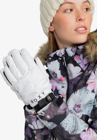 Guantes de nieve blancos con la marca Roxy que presentan una superficie texturizada, correa ajustable en la muñeca, y que se usan con una chaqueta de patrón floral.