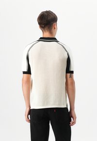Kurzärmeliges Poloshirt in Creme mit strukturiertem Strick und schwarzen Akzenten. Verfügt über einen gerippten Kragen und ein gemustertes Steppmuster entlang der Schultern.