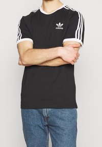 Svart bomullst-shirt med vita accenter, som har tre vita ränder på varje ärm och en liten adidas-logotyp på bröstet.