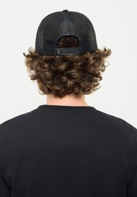 Gorra de malla negra con visera curvada, cierre a presión ajustable y una textura intrincada, llevada sobre cabello rizado castaño. Se ve una camiseta negra sencilla.