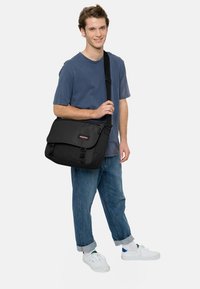 Sac messager noir avec rabat, quincaillerie en plastique, bandoulière ajustable, posé à côté d'une personne portant une chemise bleue et un jean.