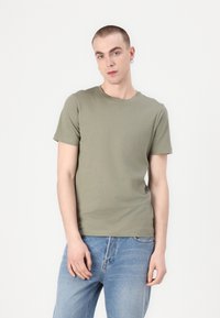 Ελαιόχρωμο βαμβακερό T-shirt με στρογγυλή λαιμόκοψη και κοντά μανίκια, σε συνδυασμό με ανοιχτό μπλε τζιν παντελόνι. Απλός σχεδιασμός, χωρίς μοτίβα ή επιχρώσεις.