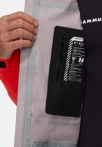 Schwarze Reißverschlusstasche auf grauem Innenfutter, Teil einer roten Jacke. Die Tasche enthält Sicherheitsanweisungen und Notfallkontakte in Weiß gedruckt.
