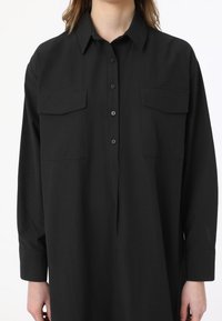 Chemise noire à manches longues avec boutons sur le devant, col et deux poches poitrine. Fabriquée dans un tissu lisse avec une coupe décontractée, elle est dotée de manches à poignets.