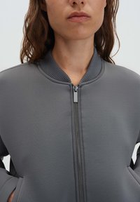 Giacca bomber grigia con collo a coste e chiusura con zip. Tessuto morbido e strutturato con tasche laterali, design minimalista e senza motivi visibili.
