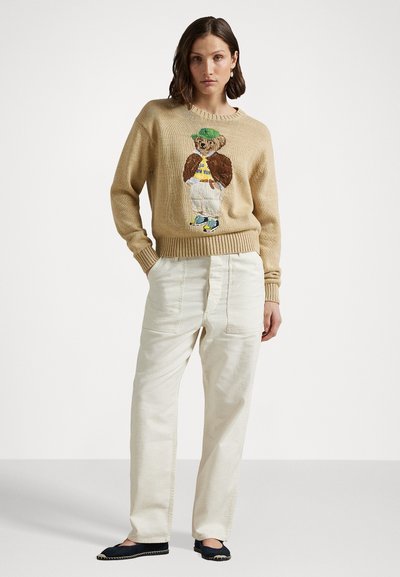 Beige gebreide trui met een geborduurd berenontwerp, gecombineerd met off-white wijde broek en marineblauwe instappers.