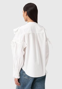 Kvinna med långt mörkt hår som bär en vit blus med volanger och puffärmar samt blå denimjeans, vänd bort mot en enfärgad bakgrund.