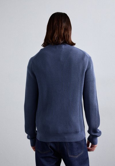 Ριμπέ μπλε πουλόβερ με μακριά μανίκια, λαιμόκοψη τύπου crew και υφή υφάσματος; εφαρμοστό στη μέση και στους καρπούς, φορεμένο πάνω από σκούρο τζιν.