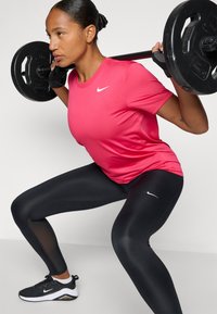 Rózsaszín sportpóló Nike logóval, fekete leggings hálós részletekkel és fekete cipő fehér díszítéssel, guggoló gyakorlat bemutatása.