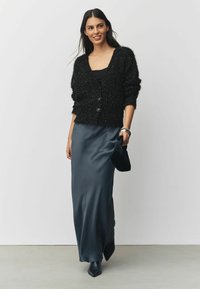 Black fluffy cardigan with a deep V-neck, two buttons, paired with a dark satin maxi skirt and black heeled boots, holding a black clutch.