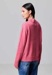 Maglione rosa a maglia con collo alto a coste, spalle scese e polsini a coste; abbinato a jeans blu in denim. Texture morbida, design casual.