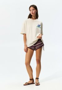 Cream t-shirt featuring green and blue shell graphic, paired with multicolored striped shorts and brown flat sandals. Casual beachwear outfit.