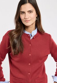 Cardigan rosso lavorato a maglia con scollo rotondo, chiusura a bottoni e due tasche frontali con dettagli a coste. Indossato sopra una camicia collare azzurra.
