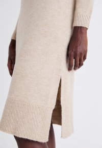 Gros plan d'une personne portant une robe en tricot beige avec une fente latérale, montrant la texture et l'ourlet côtelé, les mains détendues le long du corps.