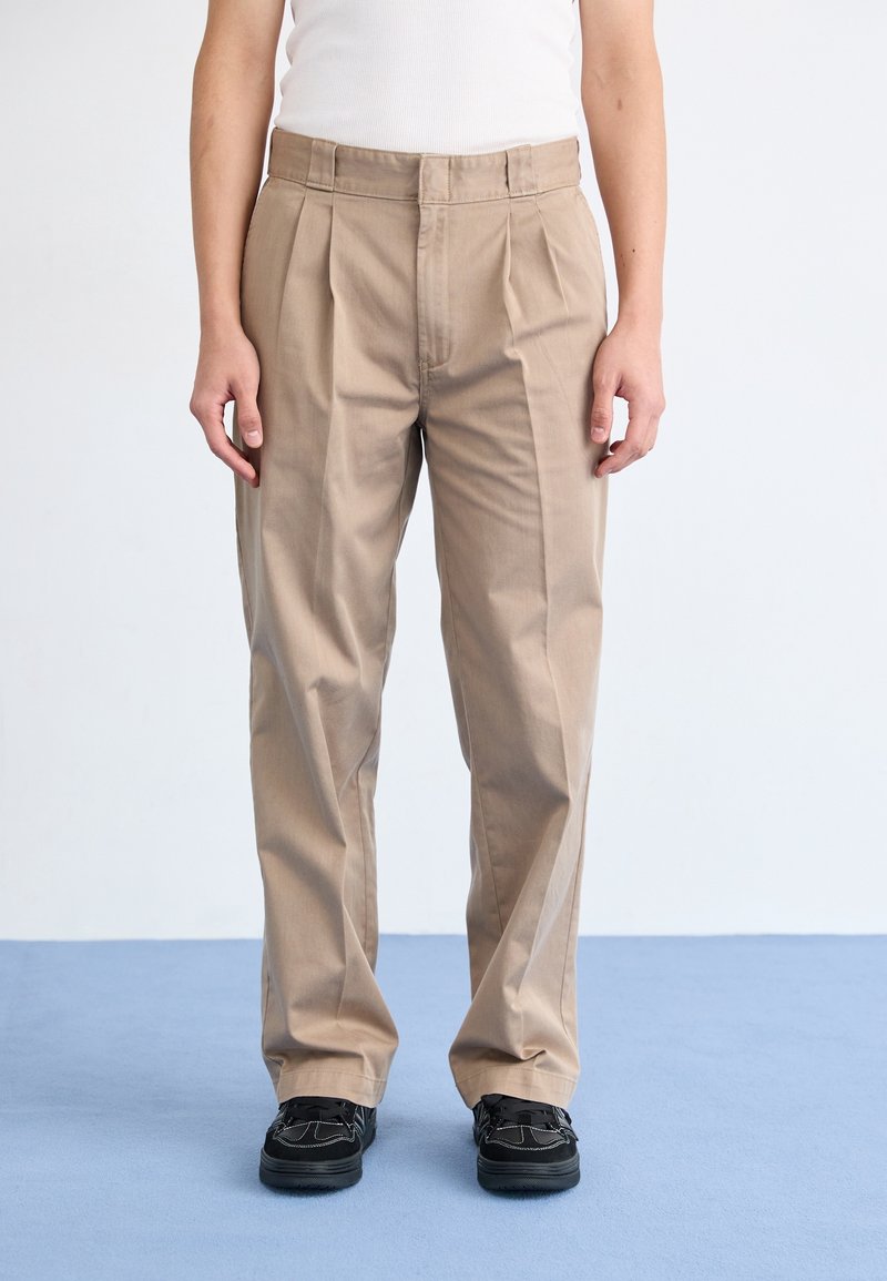 Béžové kalhoty s plisováním, vyrobené z lehké tkaniny, s přizpůsobeným střihem, rovnými nohavicemi a hladkou texturou. Černé boty doplňují vzhled.