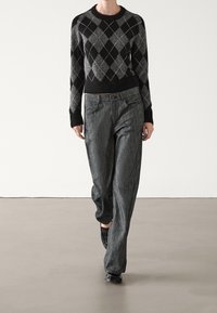 Un pull en argyle noir et gris avec un col rond, associé à un pantalon gris texturé discret et des chaussures noires.