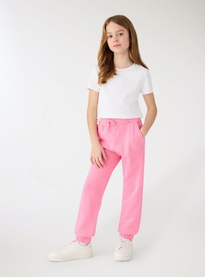 Ragazza in piedi con una mano in tasca che indossa una camicia bianca a maniche corte, pantaloni della tuta rosa e sneakers bianche su sfondo semplice.