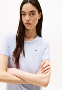 Light blue ribbed short-sleeve top with a round neckline and a small logo on the chest. The model has long dark hair, arms crossed.