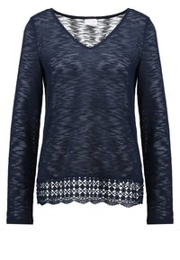 Marinblus med lång ärm och V-ringning, tillverkad av mjukt, texturerat tyg, med en spetsdetalj vid fållen som har cirkulära mönster.