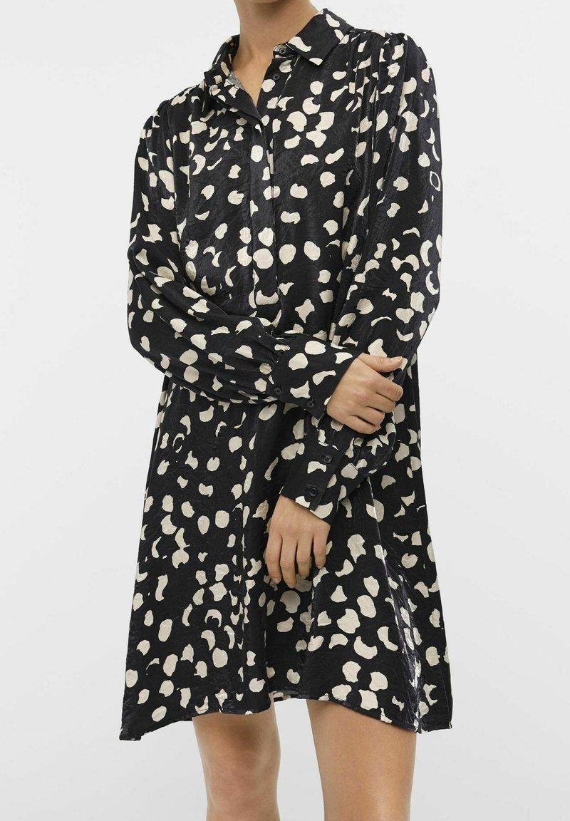 Robe noire à pois blancs irréguliers, dotée d'un col pointu et d'une fermeture à boutons sur le devant. Manches longues avec poignets à boutons. Tissu doux et texturé.