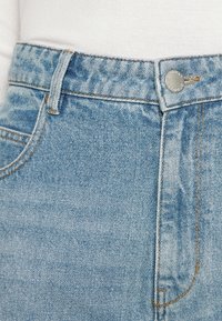 Detailní záběr na pás světle modrých džín s kovovým knoflíkem, poutkem na opasek a částí švu přední kapsy, nošených s bílým topem.
