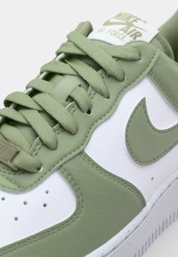 Nike Air Force 1 sneakers har en hvid læderbund med grønne accenter, struktureret gummisål og tykke snørebånd, der viser subtile syninger.