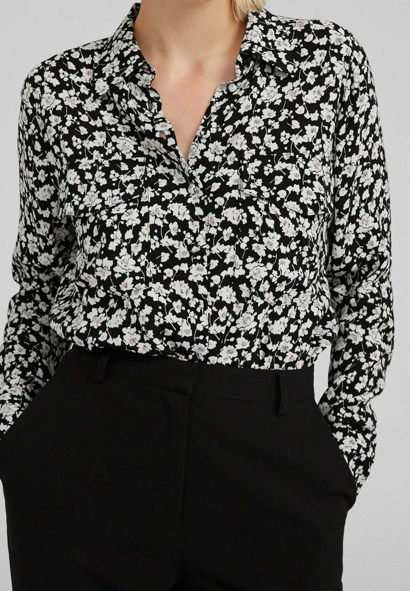 Blouse à fleurs noire et blanche à manches longues rentrée dans un pantalon noir taille haute, mains dans les poches, visible du cou aux hanches.
