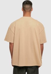 Tricou oversized bej cu mânecă scurtă, realizat din bumbac, având un gât rotund și o textură fină. Vederea din spate subliniază croiala lejeră și detaliile cusăturilor.