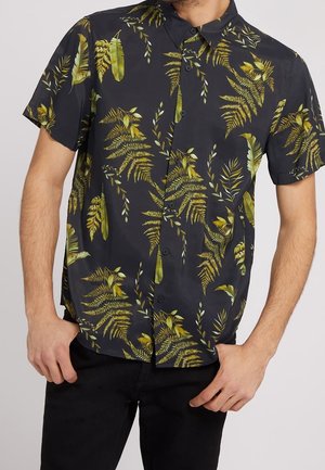 Homme portant une chemise noire à manches courtes avec un motif de feuilles tropicales jaunes et vertes, associée à un pantalon noir.