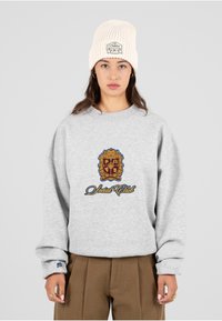 Jonge vrouw met een crèmekleurige gebreide beanie en een grijs sweatshirt met een geel en blauw "Social Club" embleem, gecombineerd met een bruine broek.
