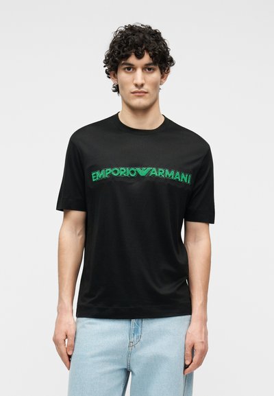 Νέος με κατσαρά μαλλιά που φοράει μαύρο t-shirt με πράσινο λογότυπο «EMPORIO ARMANI» και ανοιχτόχρωμα μπλε τζιν, στέκεται μπροστά σε απλό φωτεινό φόντο.