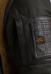 Bruine leren patch op een donkergroene stoffen jas, met getextureerde stiksels en een klein label dat leest "PME LEGEND TESTED AT HIGH ALTITUDE."