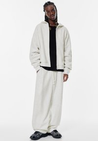Jasnoszara, oversize'owa bluza z kapturem na zamek, połączona z luźnymi, szarymi spodniami dresowymi i czarnymi sneakerami z teksturowanymi akcentami.