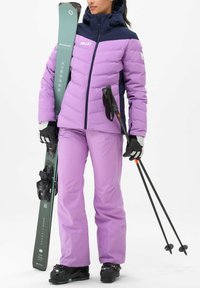 Lila isolierte Skijacke mit einem marineblauen Oberteil, passenden lila Skihosen, schwarzen Handschuhen und Skiausrüstung, einschließlich Skiern und Stöcken.