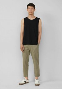 Junger Mann steht, trägt ein schwarzes ärmelloses Shirt, beige Cropped-Hosen mit Taschen und weiße Sneaker mit schwarzen Akzenten.